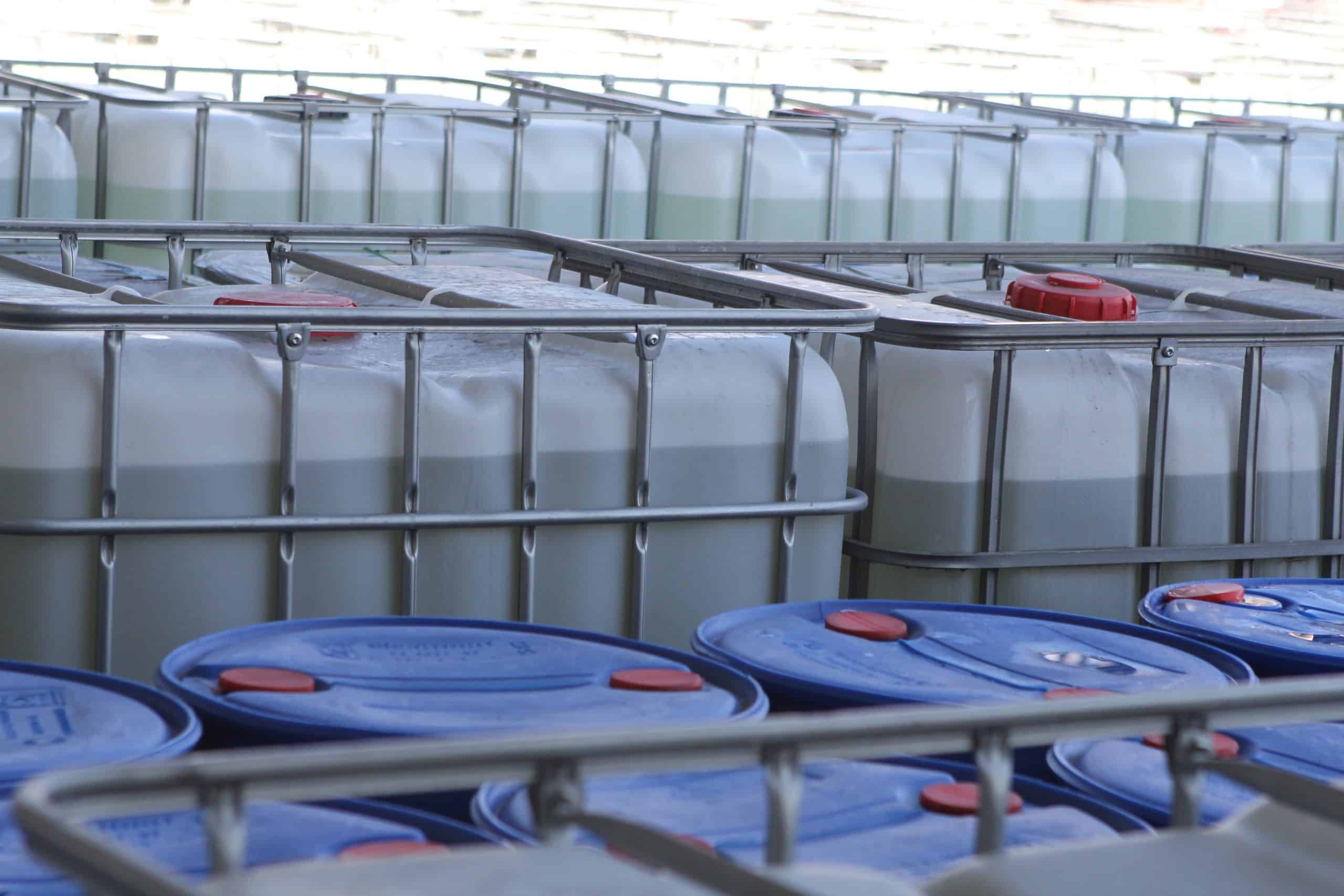 Colonial Chemical Solutions, Inc. Rows of large white industrial liquid storage containers (IBC totes) inside a facility, each partially filled with light-colored liquid and secured in metal frames, with blue barrels visible in the foreground and red caps on the containers.
